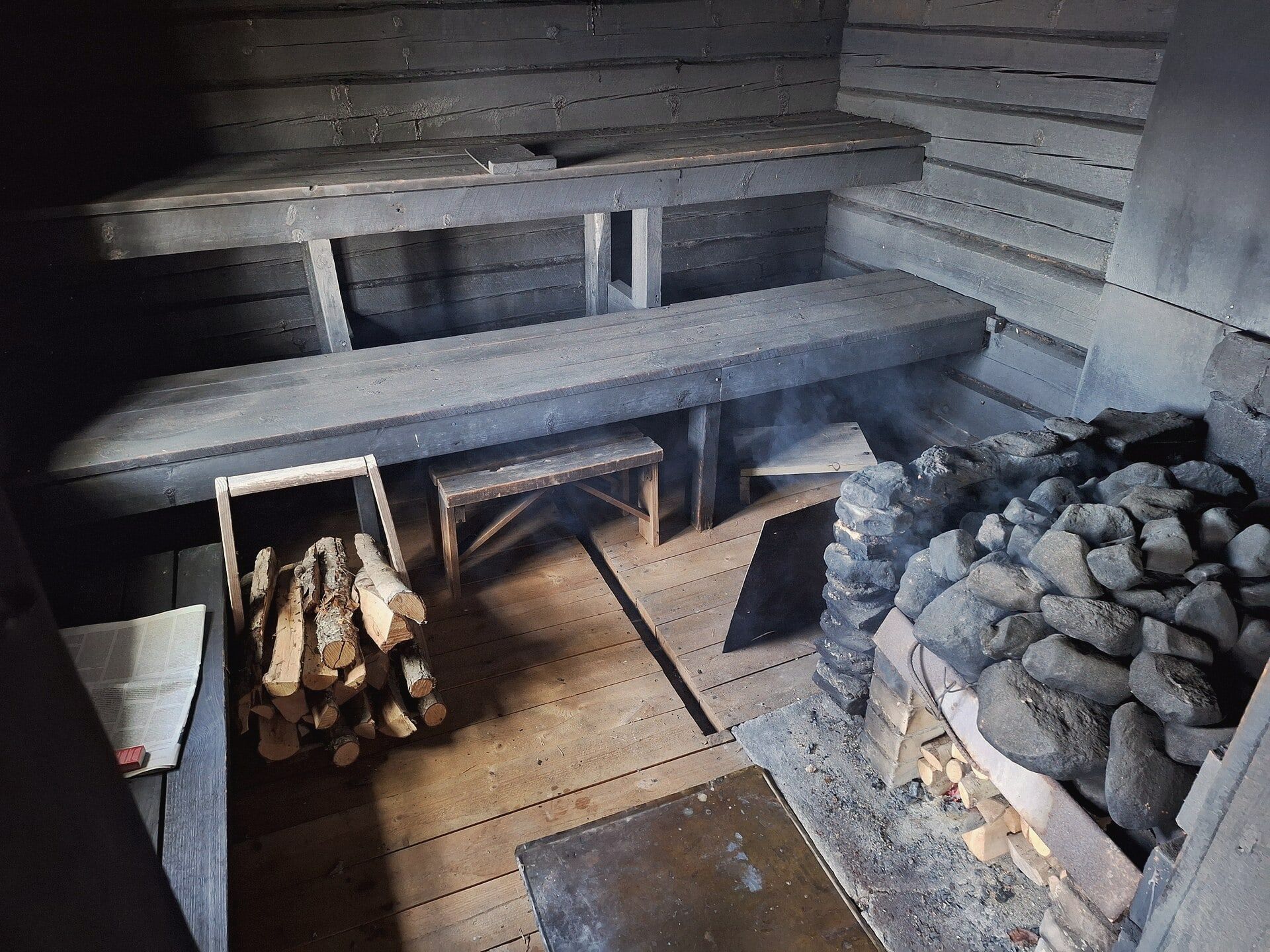 Inside a wood-heated sauna: weathered benches, a stone heater and stacked firewood.